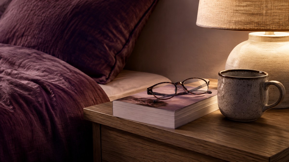 A book, glasses and ceramic cup on a bedside table.